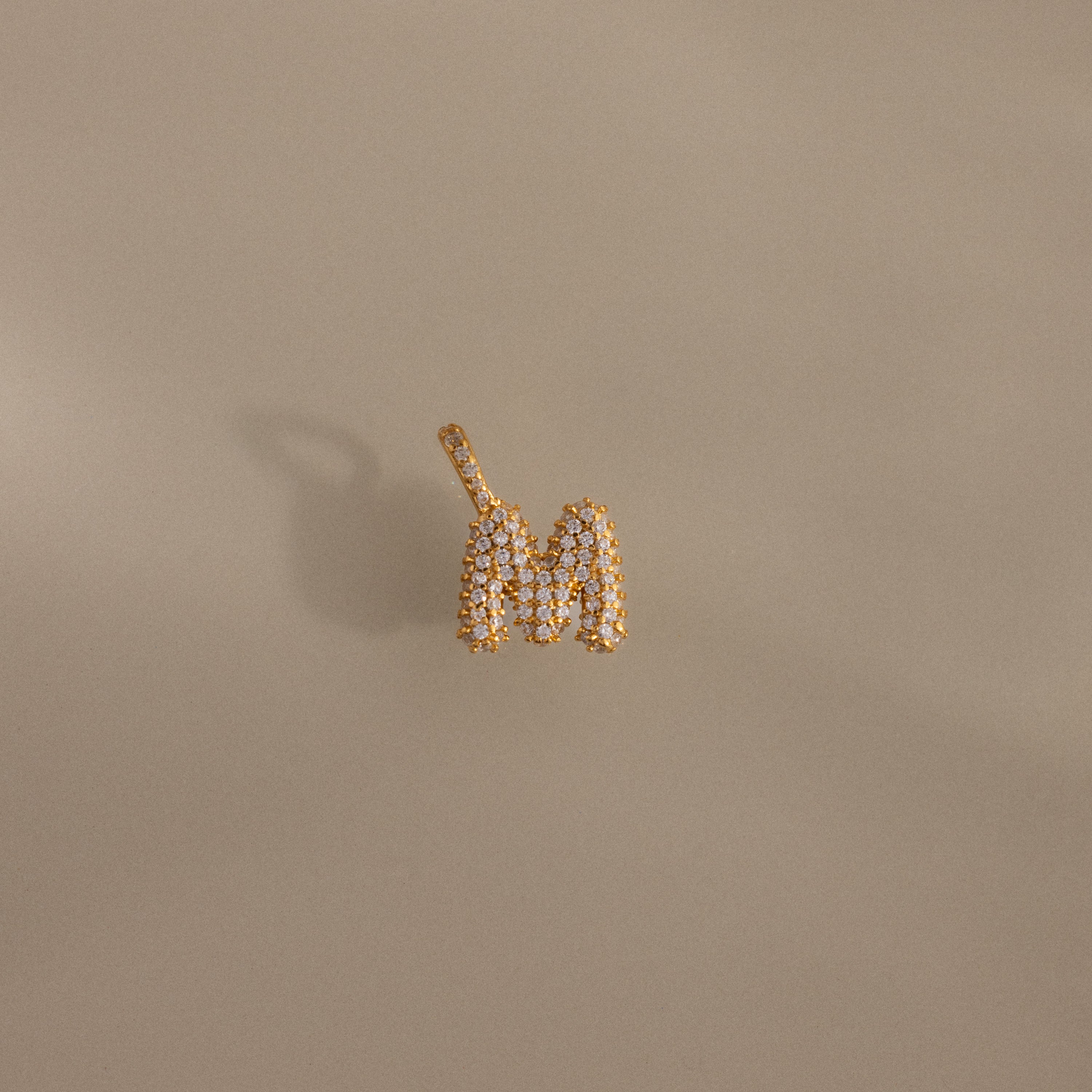 The Pave Bubble Letter Charm is a gold and diamond pendant shaped like the letter M, featuring diamond-studded lettering on a beige background.