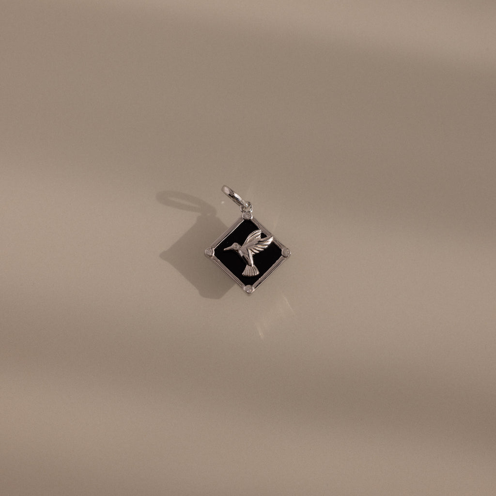 The Onyx Hummingbird Charm in Silver rests on a smooth beige surface with soft shadows—a stylish accessory that brings a unique flair to your jewelry collection.