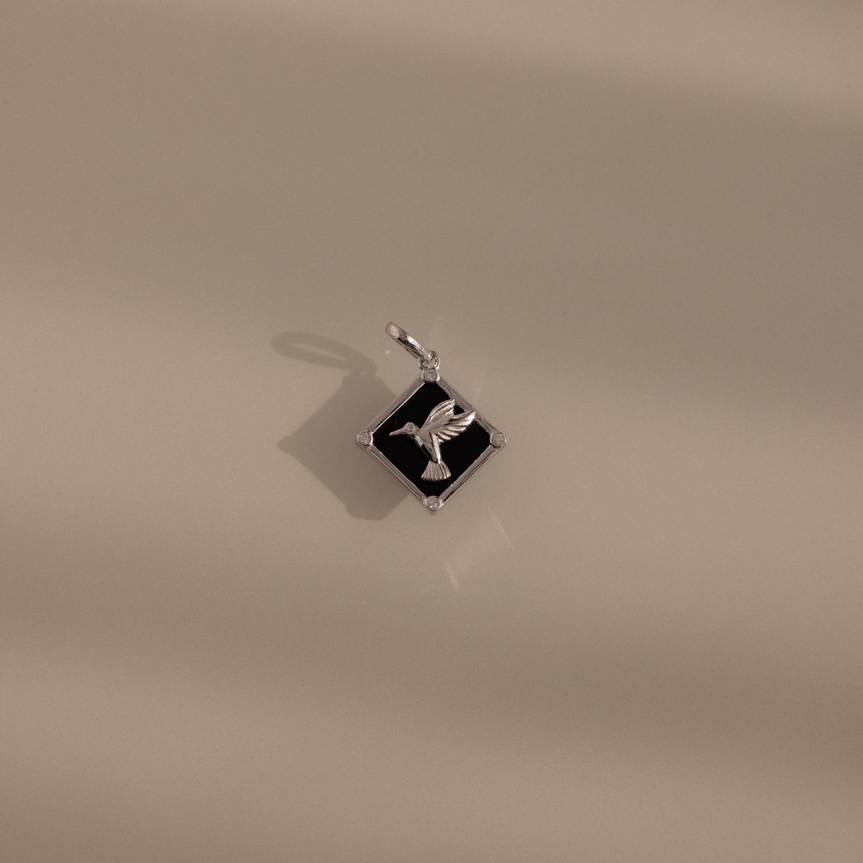 The Onyx Hummingbird Charm in Silver rests on a smooth beige surface with soft shadows—a stylish accessory that brings a unique flair to your jewelry collection.