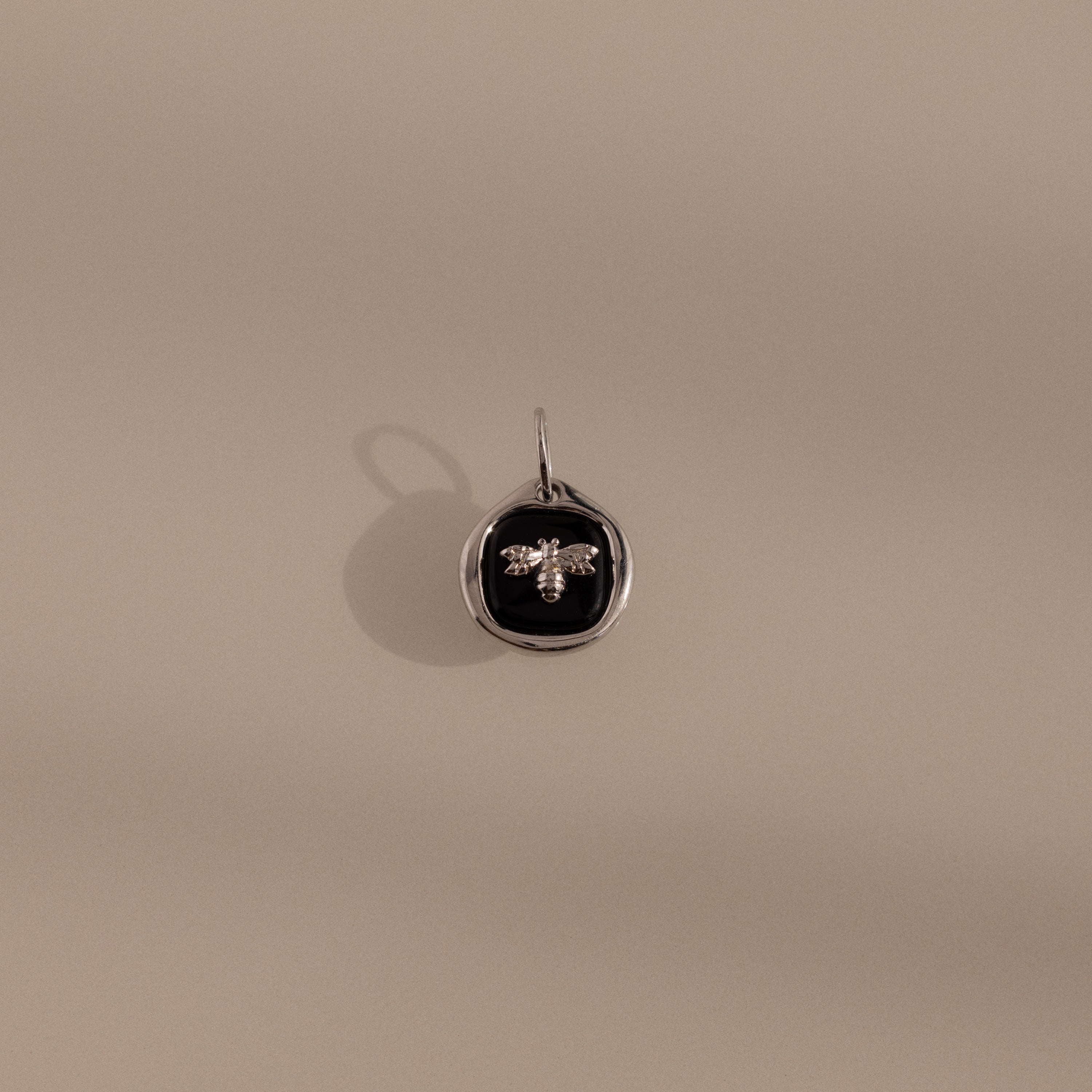 The Onyx Honeybee Charm in Silver features a raised bee on a black onyx oval, shown against a beige background—a striking accessory that effortlessly elevates your style.
