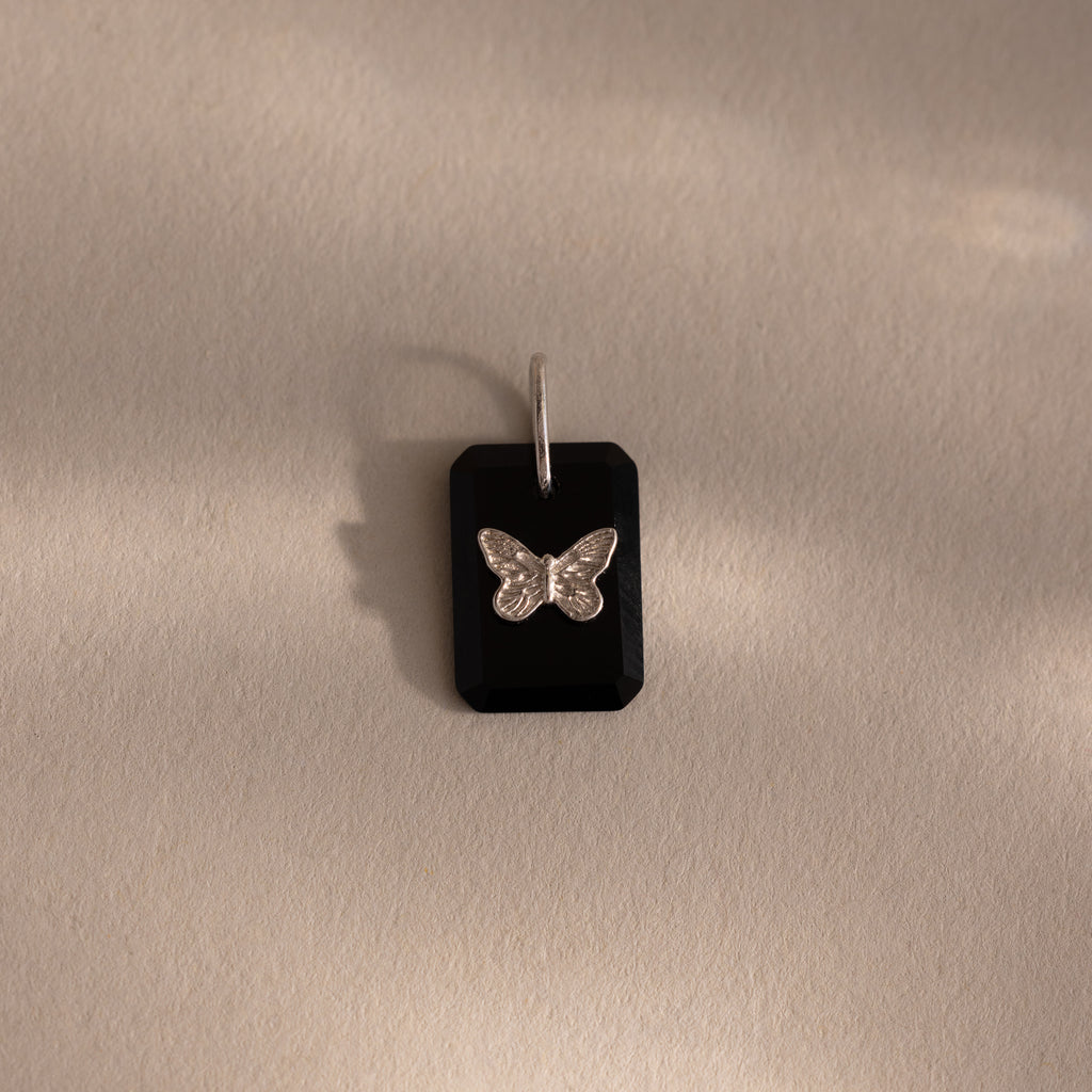 The Onyx Butterfly Charm in Silver features a black rectangular pendant with a silver butterfly design, displayed on a beige surface—an elegant addition to any jewelry collection.