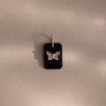 The Onyx Butterfly Charm in Silver features a black rectangular pendant with a silver butterfly design, displayed on a beige surface—an elegant addition to any jewelry collection.