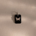 The Onyx Butterfly Charm in Silver features a black rectangular pendant with a silver butterfly design, displayed on a beige surface—an elegant addition to any jewelry collection.
