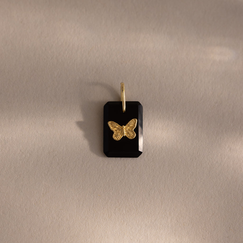 The Onyx Butterfly Charm in 18K Gold is a black rectangular pendant with a gold butterfly on a soft beige background, perfect for adding an elegant touch to any outfit.