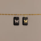 The Onyx Butterfly Charm in Silver features two black rectangular pendants with butterfly designs on a silver chain, making an elegant statement against a beige background.