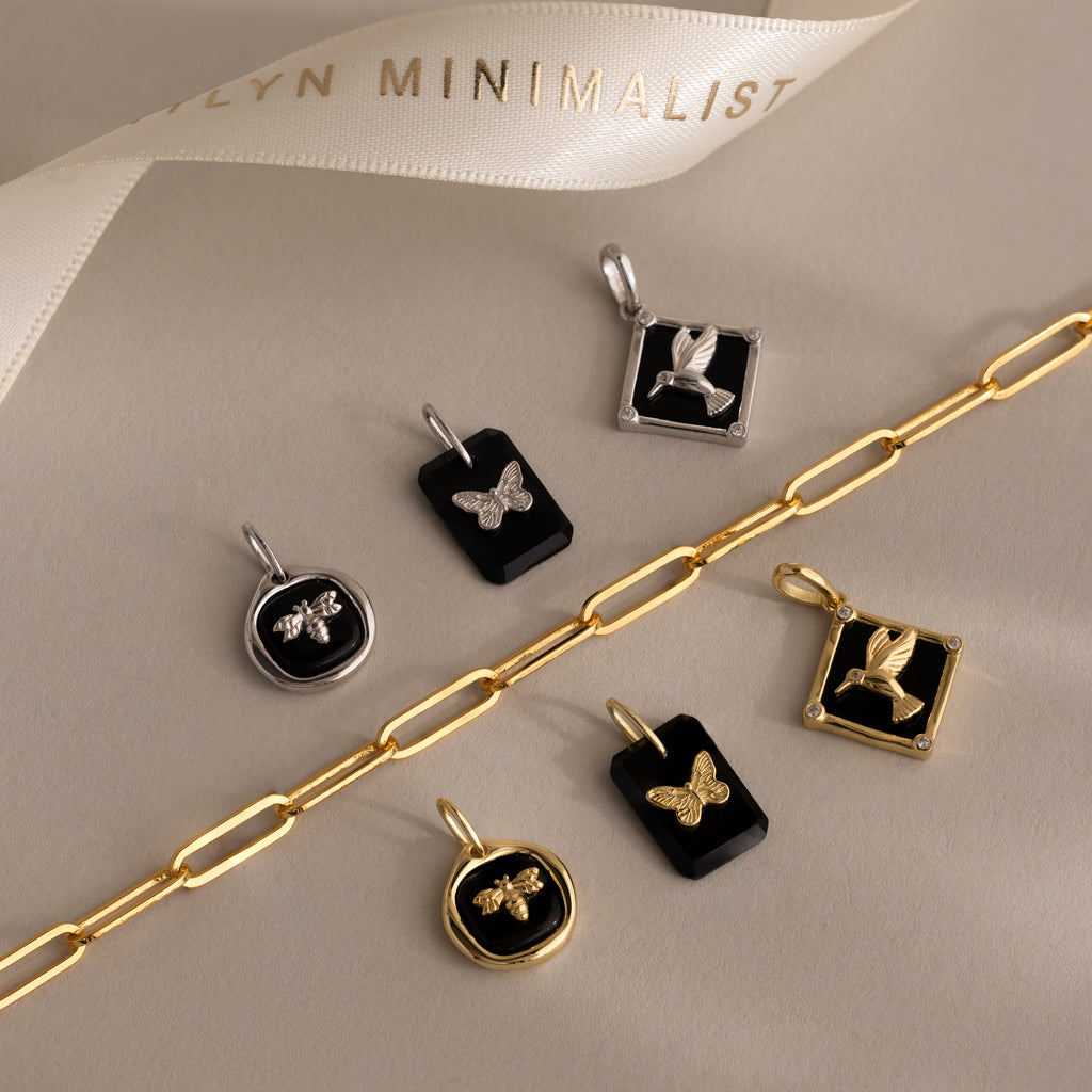 The Onyx Butterfly Charm in Silver is displayed on a beige surface with other gold and silver jewelry pieces, creating an elegant, nature-inspired collection.