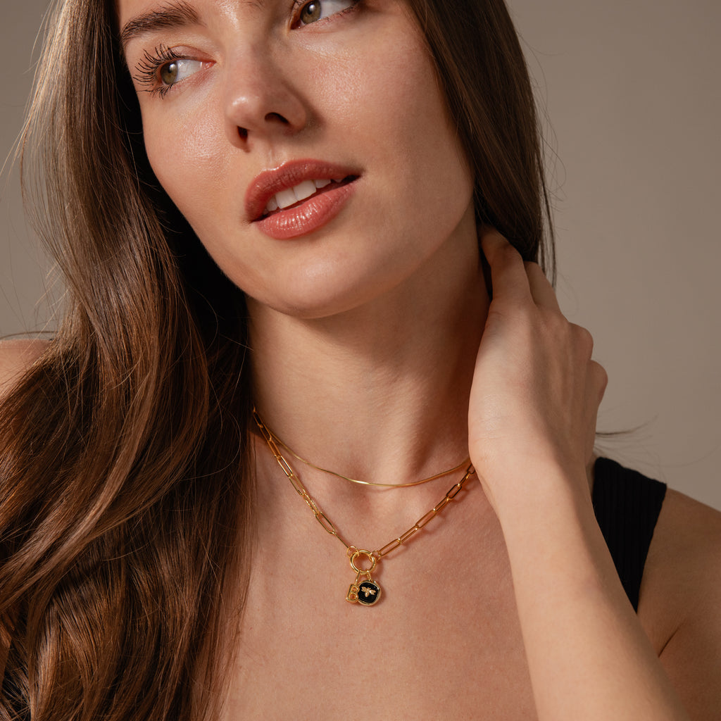 Woman with long brown hair wears a black top and layers gold necklaces, featuring the Onyx Honeybee Charm, gazing to the side with her hand on her neck for a timeless, elegant look.