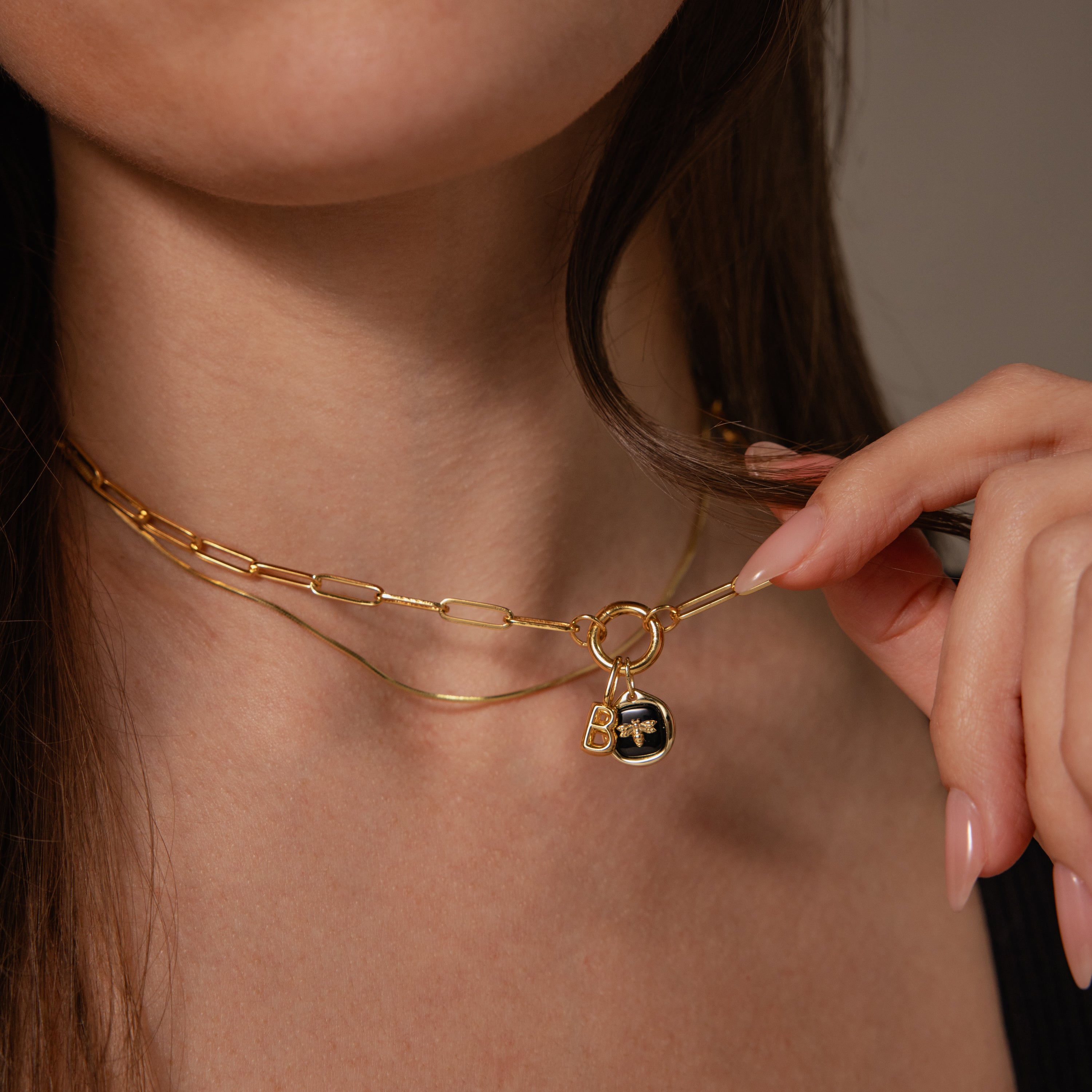 Close-up of a woman wearing layered gold chain necklaces, featuring the Onyx Honeybee Charm with a round bee pendant, delicately touching the jewelry for an elegant look.