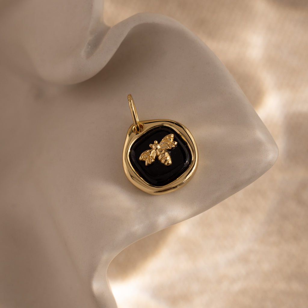 The Onyx Honeybee Charm in 18K Gold glows against a black background, displayed elegantly on a wavy cream surface.