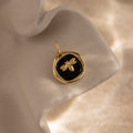 The Onyx Honeybee Charm in 18K Gold glows against a black background, displayed elegantly on a wavy cream surface.