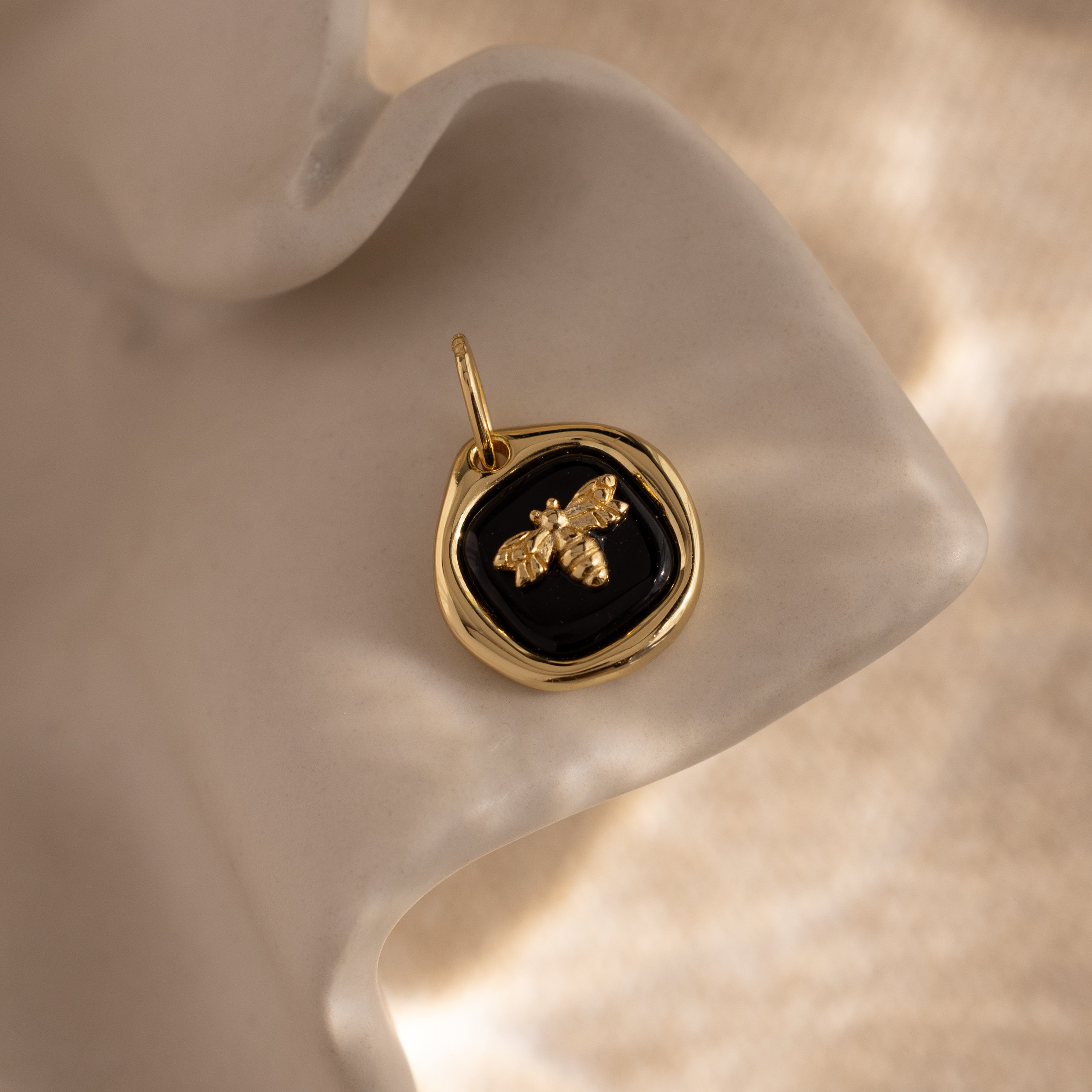 The Onyx Honeybee Charm in 18K Gold glows against a black background, displayed elegantly on a wavy cream surface.