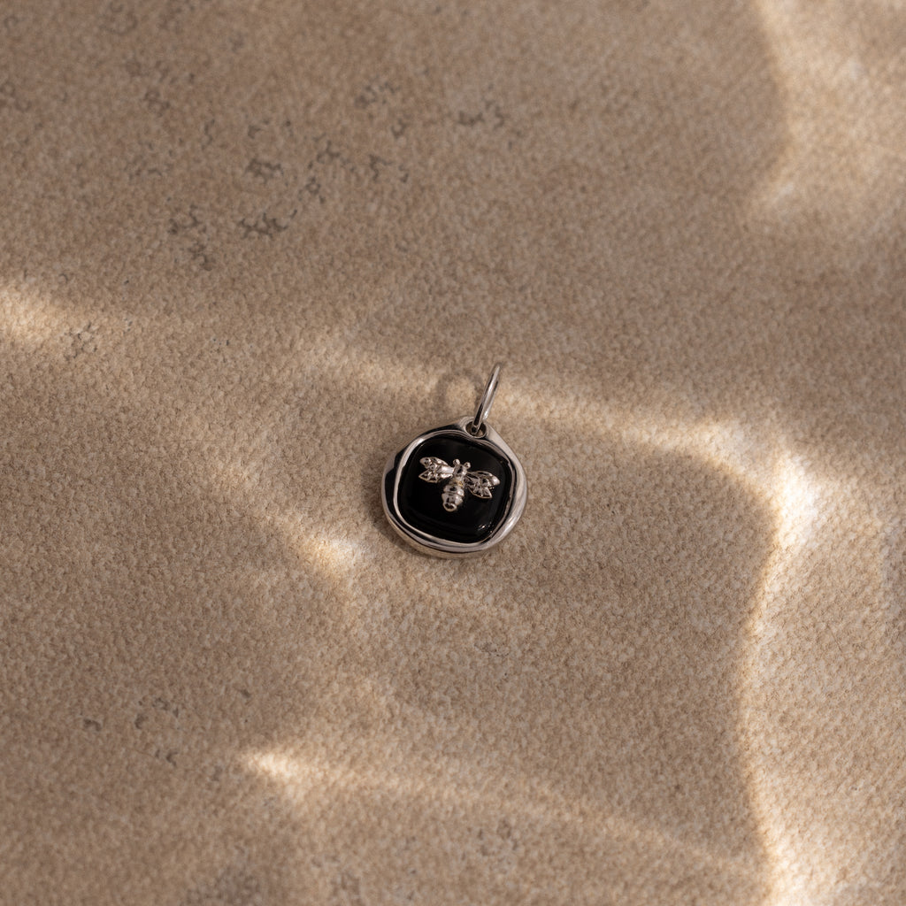 The Onyx Honeybee Charm in Silver rests on textured sand, showcased against a black background with soft sunlight.