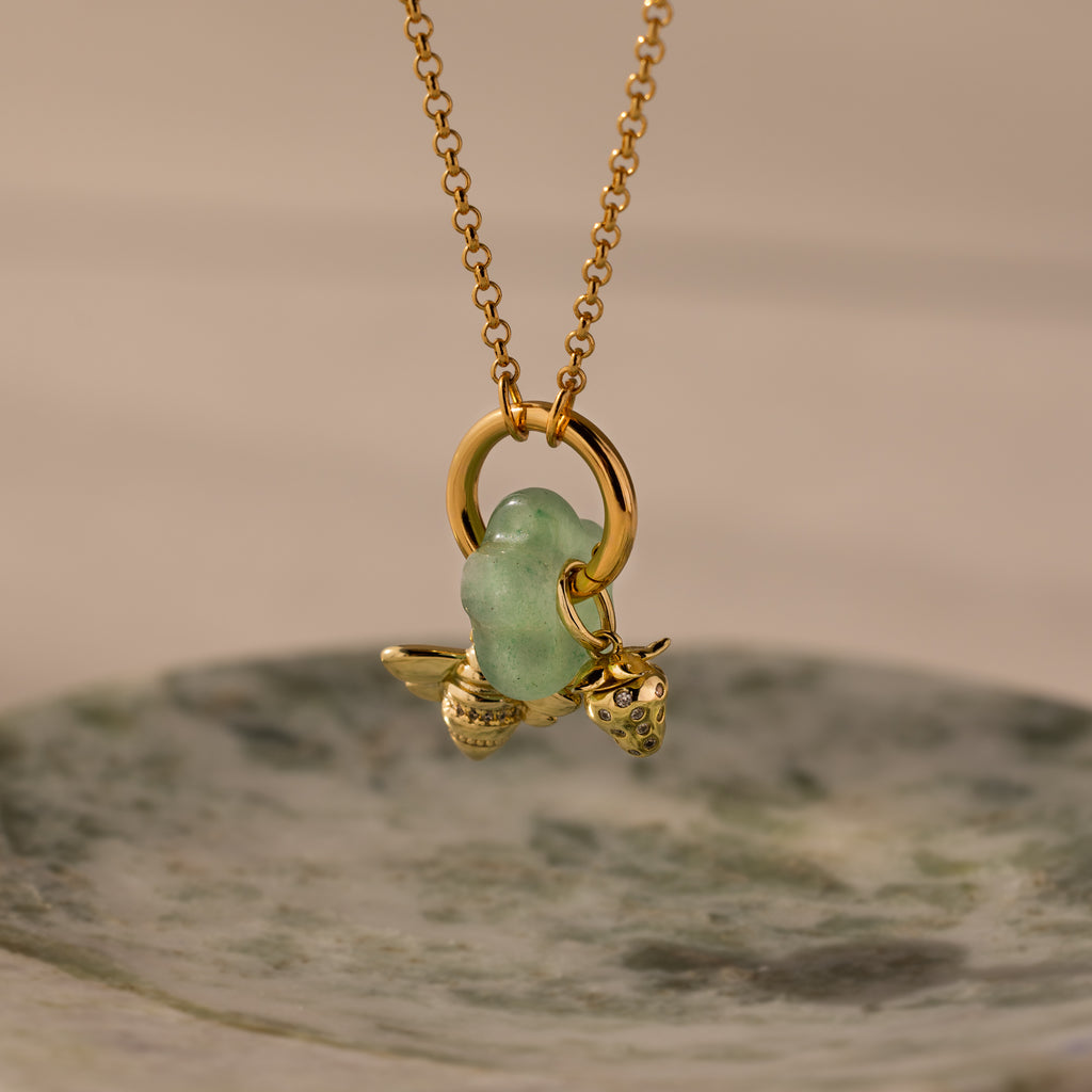 The Jade Flower Charm features a gold necklace with a green glass bead and delicate gold bee and strawberry charms, beautifully displayed on a marble dish—ideal for nature-inspired jewelry enthusiasts seeking unique accessories.