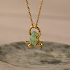 The Jade Flower Charm features a gold necklace with a green glass bead and delicate gold bee and strawberry charms, beautifully displayed on a marble dish—ideal for nature-inspired jewelry enthusiasts seeking unique accessories.