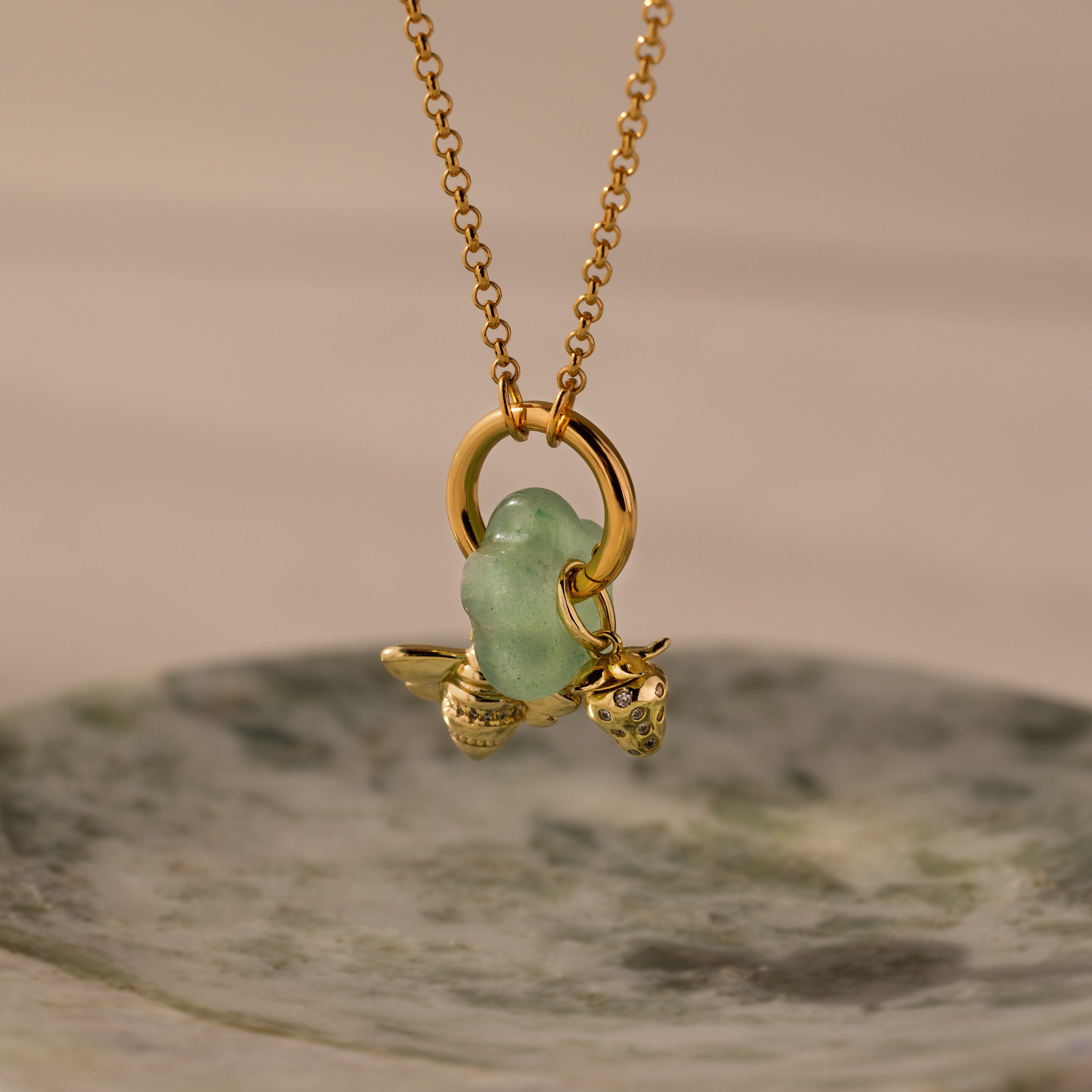 The Jade Flower Charm features a gold necklace with a green glass bead and delicate gold bee and strawberry charms, beautifully displayed on a marble dish—ideal for nature-inspired jewelry enthusiasts seeking unique accessories.