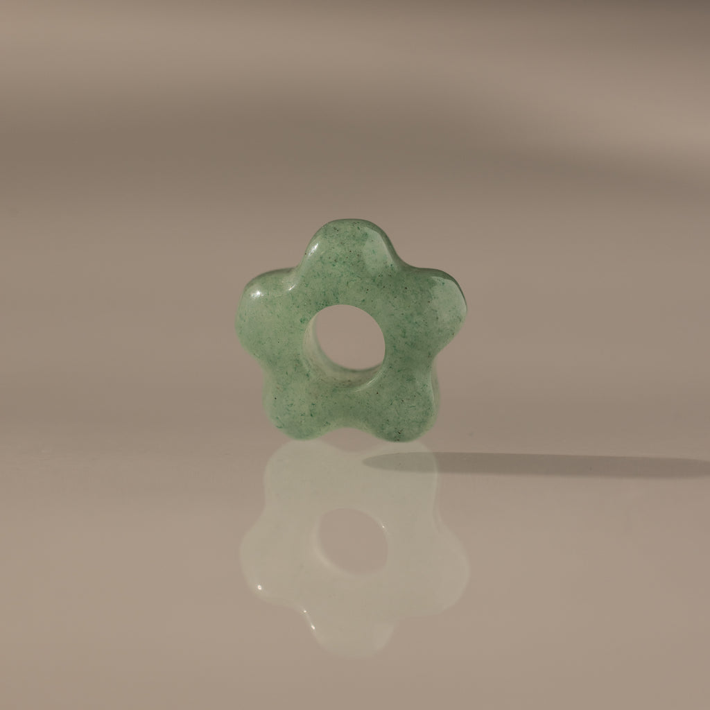 The Jade Flower Charm, a green jade bead shaped like a flower with a central hole, stands upright on a reflective surface and displays its polished elegance.