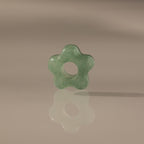 The Jade Flower Charm, a green jade bead shaped like a flower with a central hole, stands upright on a reflective surface and displays its polished elegance.