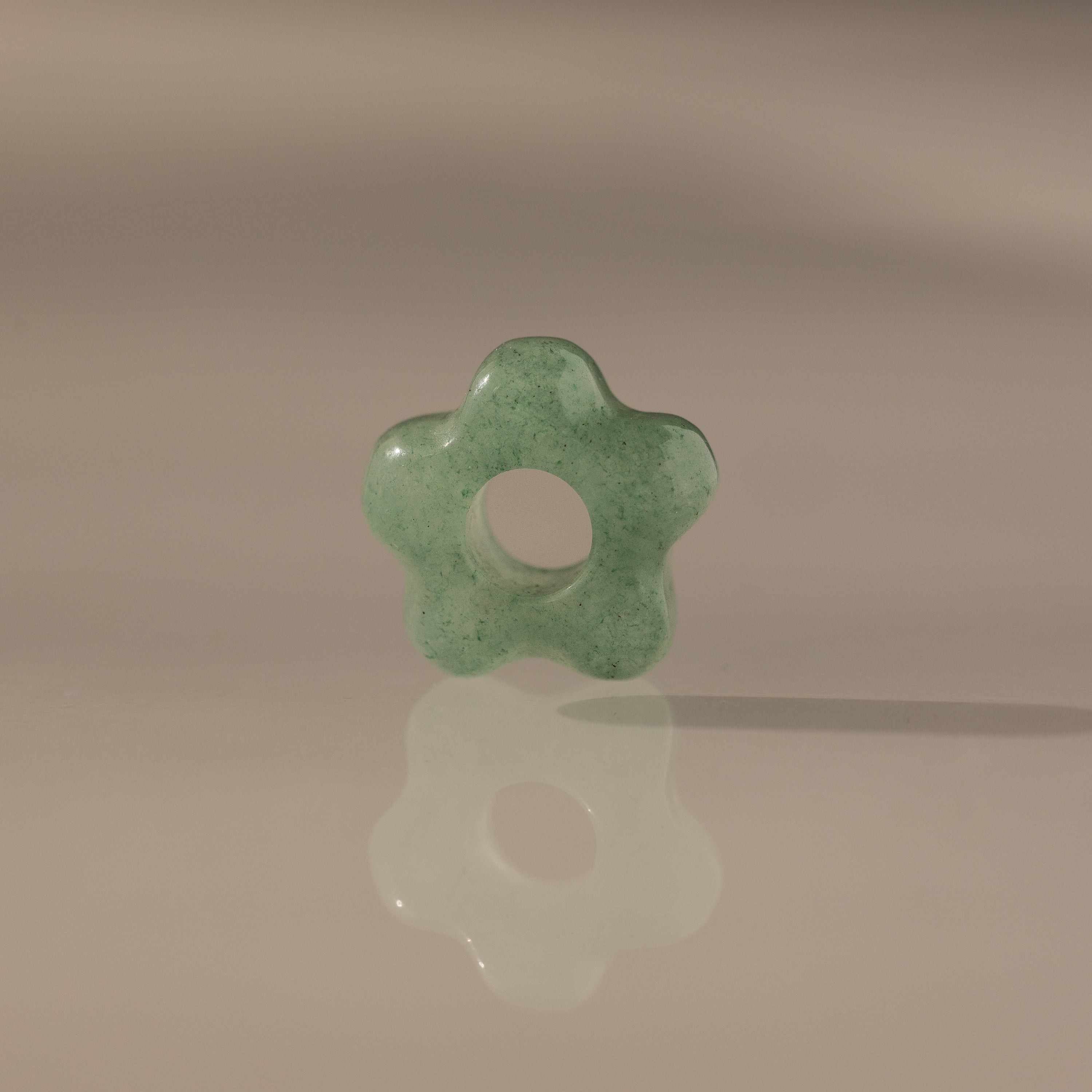 The Jade Flower Charm, a green jade bead shaped like a flower with a central hole, stands upright on a reflective surface and displays its polished elegance.