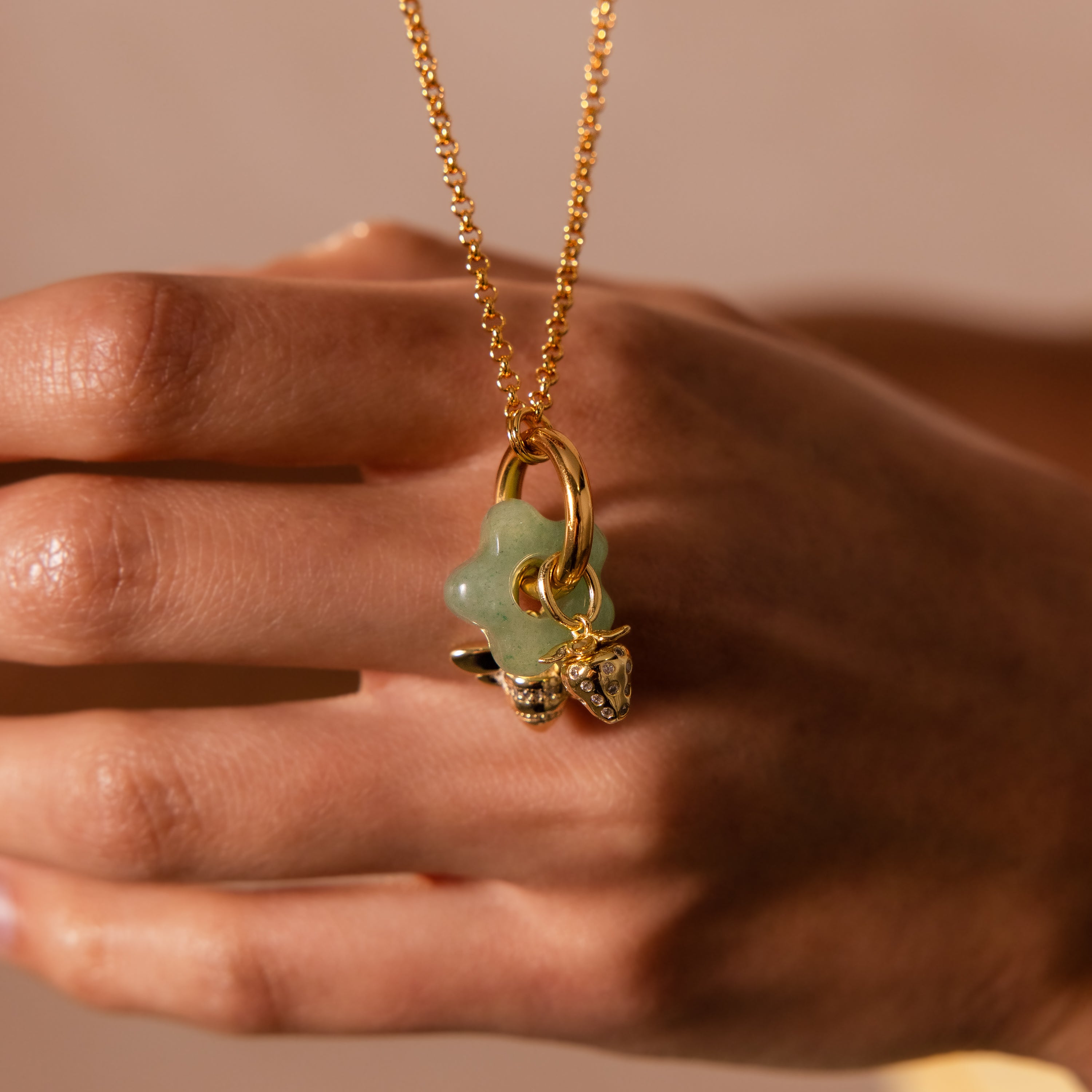 A hand holds the Jade Flower Charm—a gold necklace featuring a green flower pendant and small gold charms, making it an elegant accessory perfect for nature-inspired style.