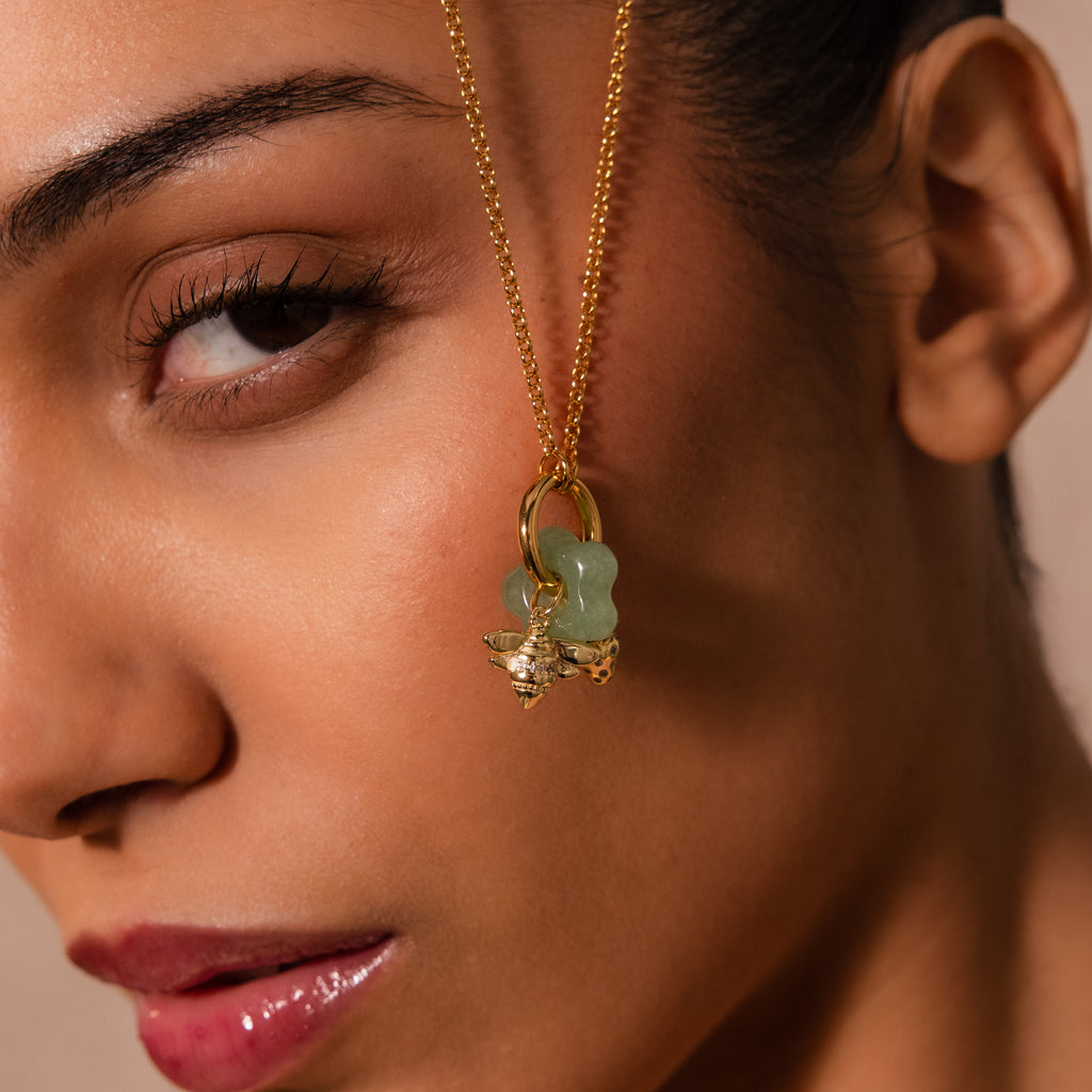 A close-up of a woman's face features the Jade Flower Charm, a gold necklace with a green pendant held near her temple, highlighting elegant green stone jewelry.