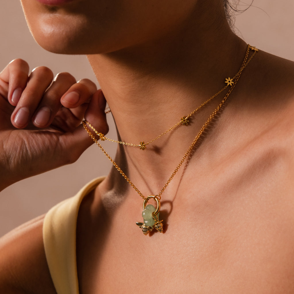 A woman wears layered gold necklaces featuring the Jade Flower Charm, a green stone pendant. She holds one chain near her neck, showcasing an elegant stack with a vibrant gemstone for effortless, eye-catching style.