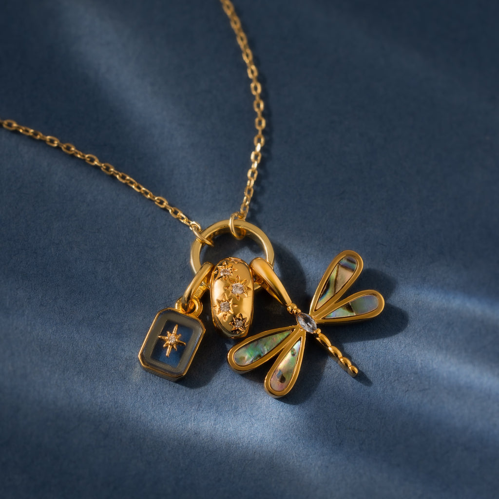 18K gold necklace featuring the Sprinkle Diamond Dome Charm, paired with a starburst stones pendant, an oval with diamonds, and a dragonfly charm on blue.