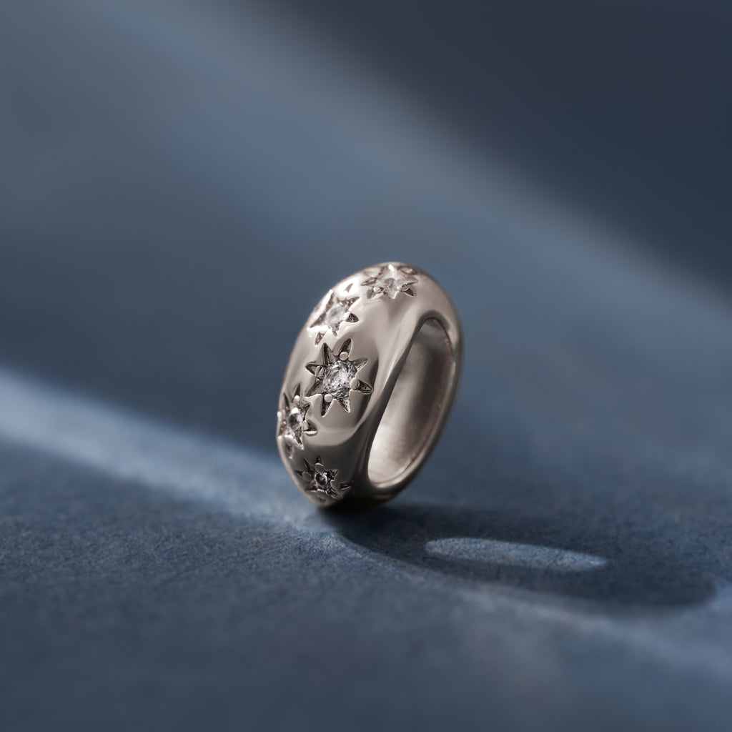 The Sprinkle Diamond Dome Charm, a silver ring with engraved star patterns and starburst-set small diamonds, rests on a smooth blue surface.