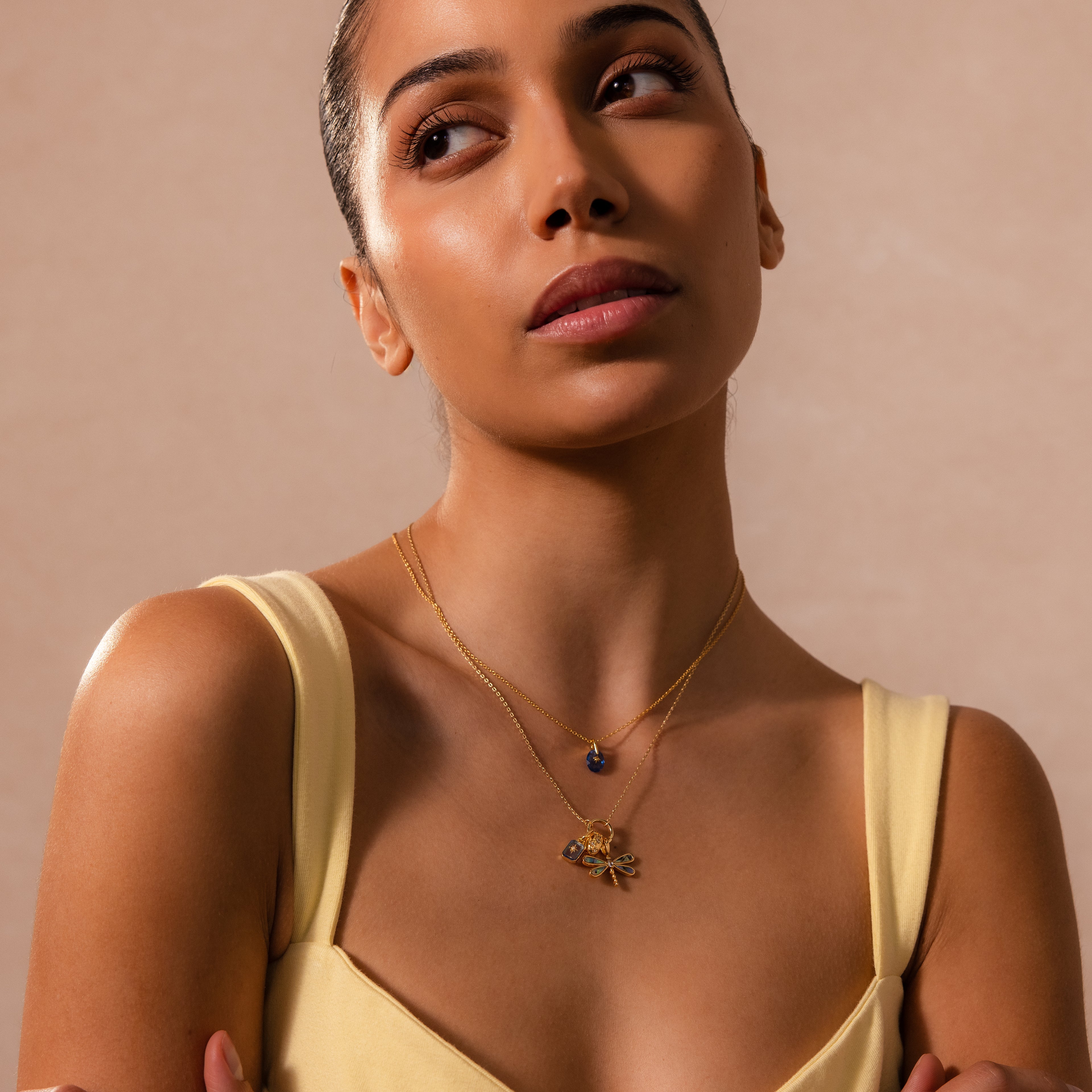 A woman in a yellow top wears two layered gold necklaces, featuring the Sprinkle Diamond Dome Charm, as she gazes slightly upward against a beige background.