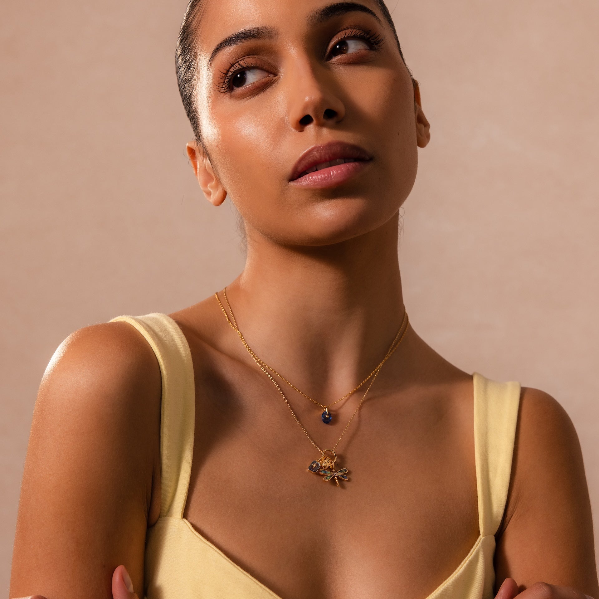 A woman in a yellow dress layers gold necklaces, featuring the Abalone Dragonfly Charm in 18K Gold, and looks upward against a neutral backdrop. The charm adds a subtle, nature-inspired accent to her sophisticated look.