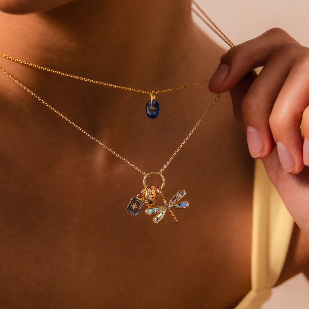 Woman wearing two gold necklaces: one with a blue pendant, and one featuring a charm stack that includes the Sprinkle Diamond Dome Charm and a dragonfly.