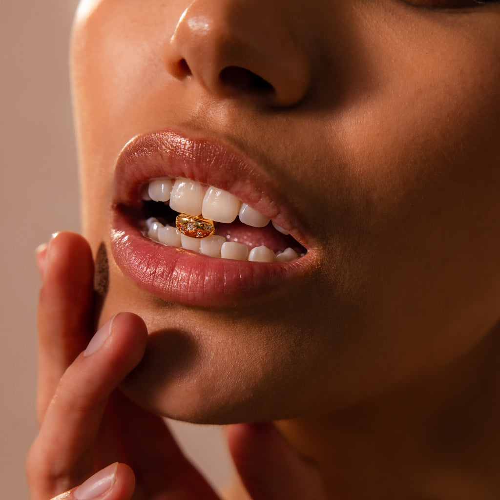 Close-up of lips and teeth adorned with the Sprinkle Diamond Dome Charm in 18K Gold, featuring starburst-set stones, as the person gently touches their face.