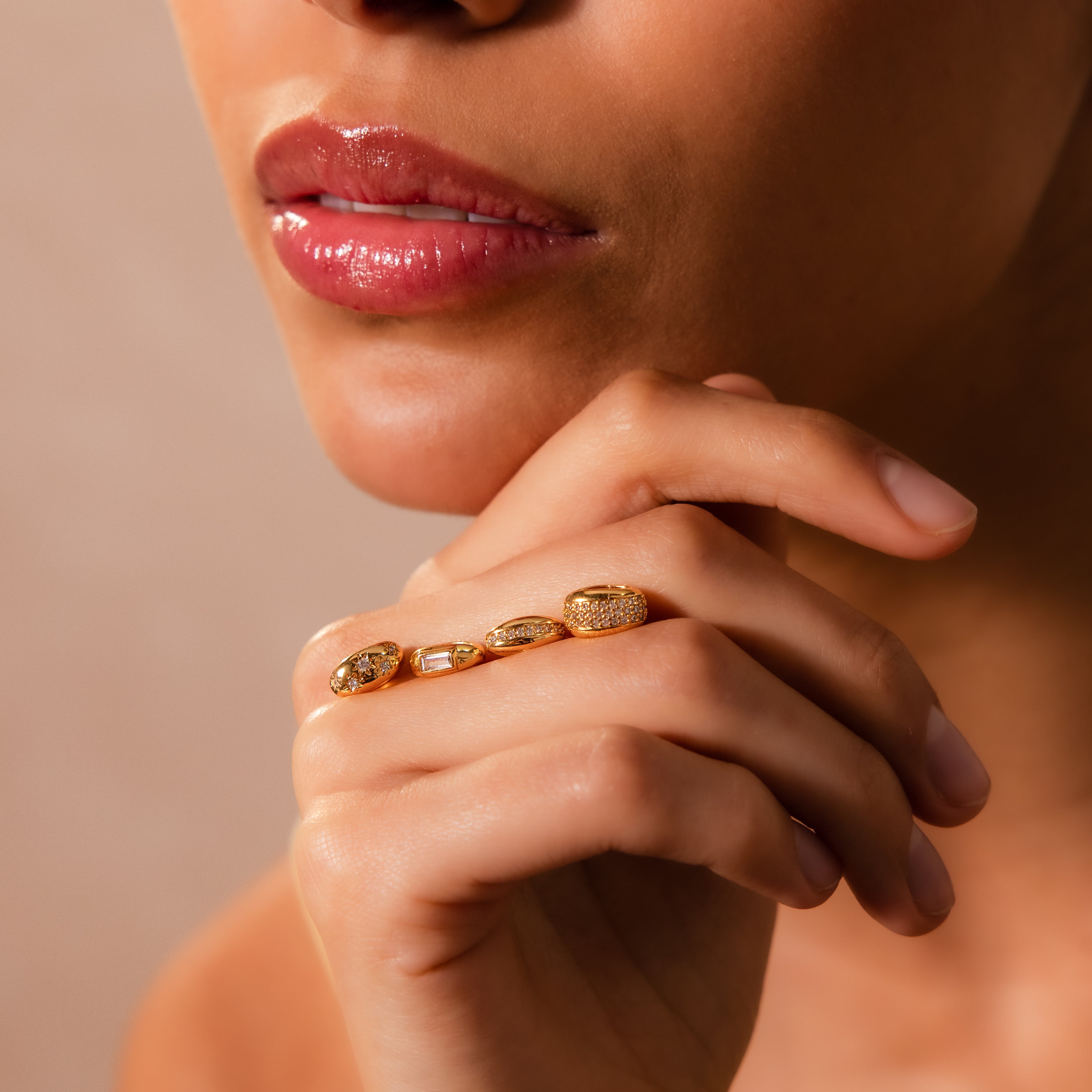 A woman's hand adorned with gold rings and the Sprinkle Diamond Dome Charm rests near her lips, highlighting her glossy makeup and smooth, radiant skin.