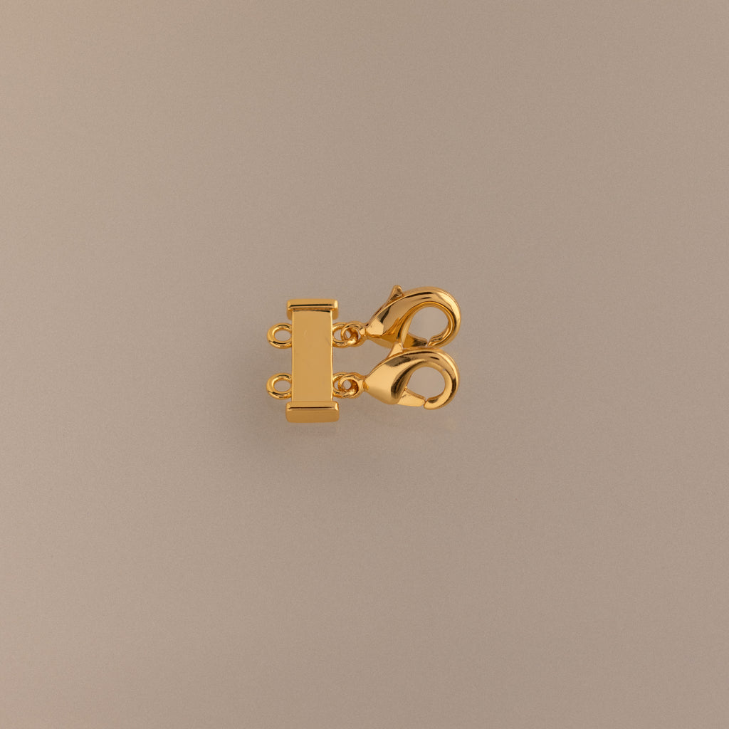 The Double Layering Clasp in 18K Gold features a triple-strand design with two lobster claw closures, ideal for layering necklaces. Displayed on a neutral beige background.