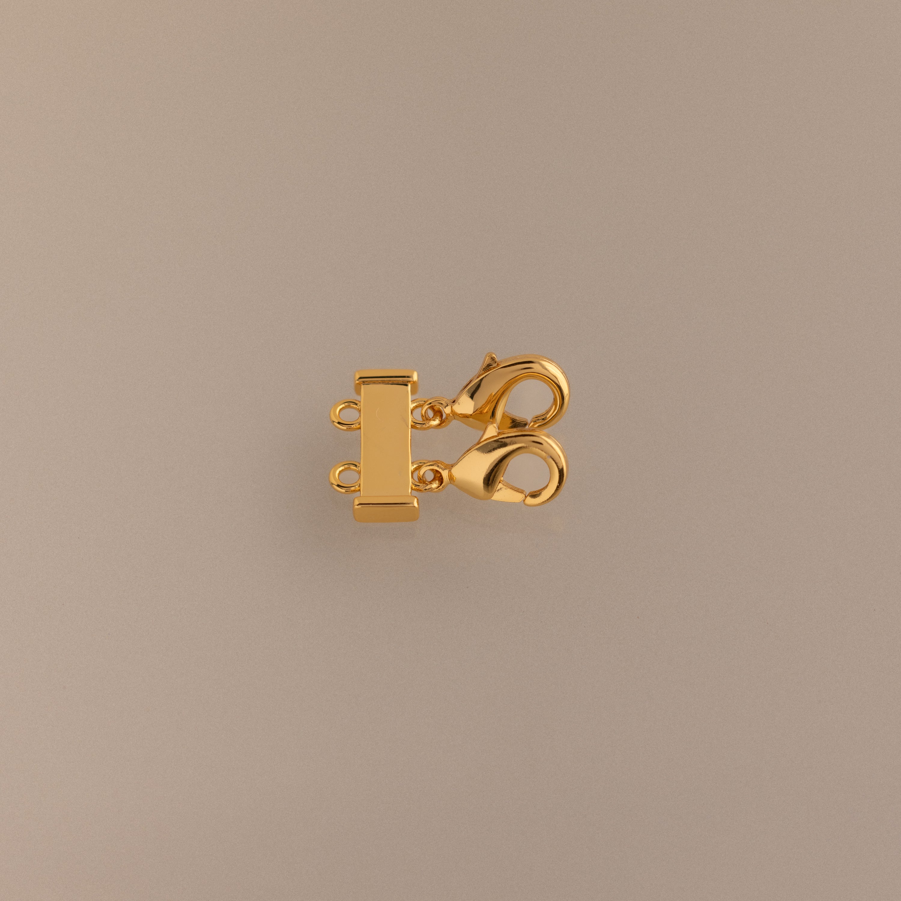 The Double Layering Clasp is a gold multi-strand jewelry clasp with two lobster clasps and three connector rings, perfect for necklace layering and preventing tangling—shown on a beige background.