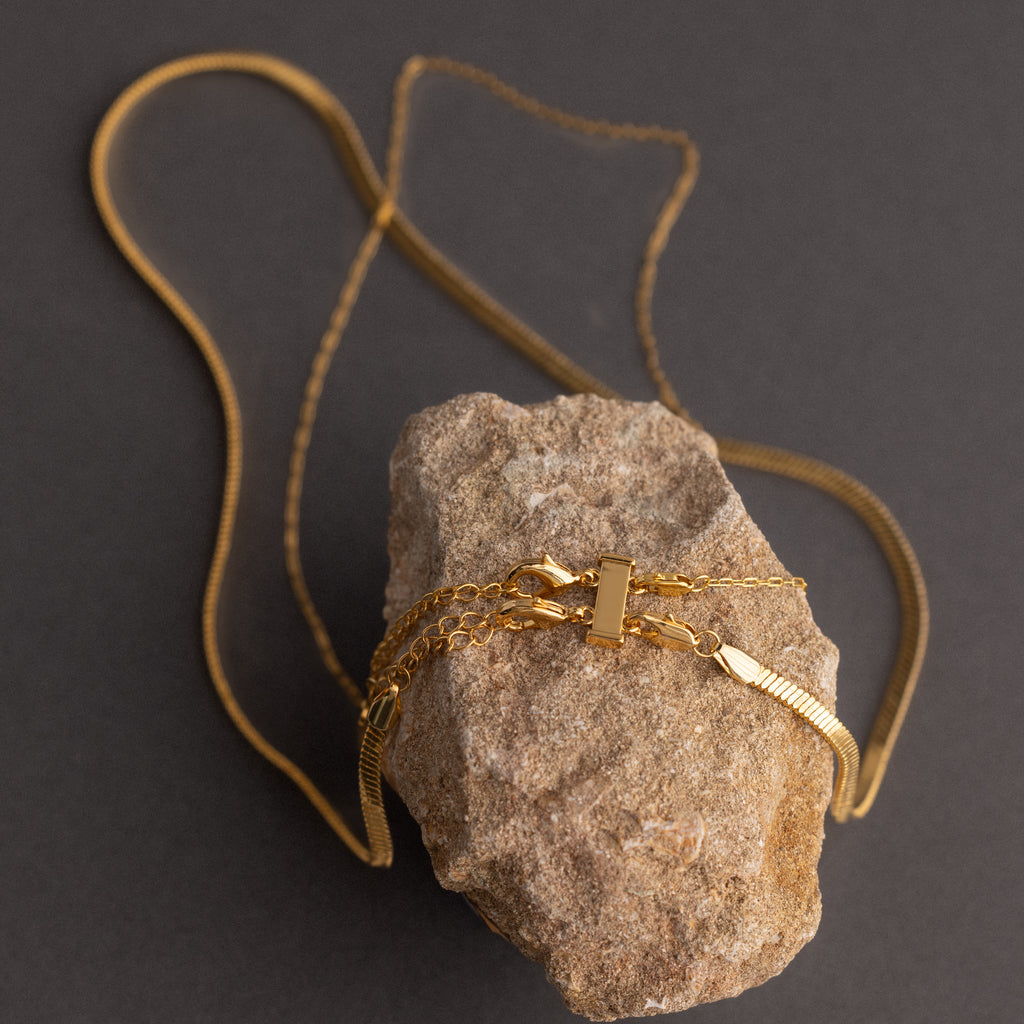 The Double Layering Clasp, a gold necklace accessory, wraps around a rough beige stone on a dark background, showcasing anti-tangle features for effortless necklace layering.