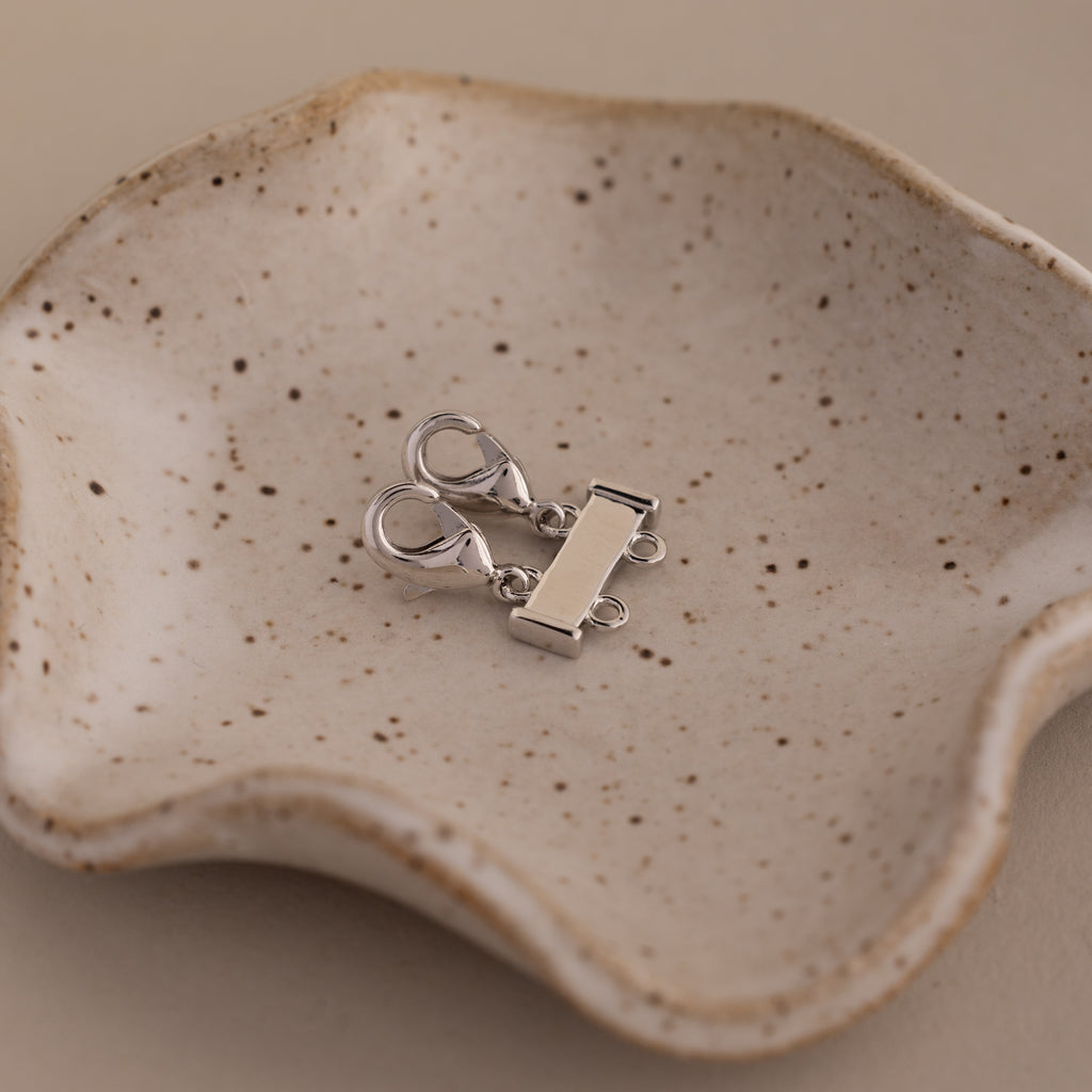 The Double Layering Clasp in Silver rests on a speckled ceramic dish with wavy edges, perfect for layering necklaces, set against a beige background.