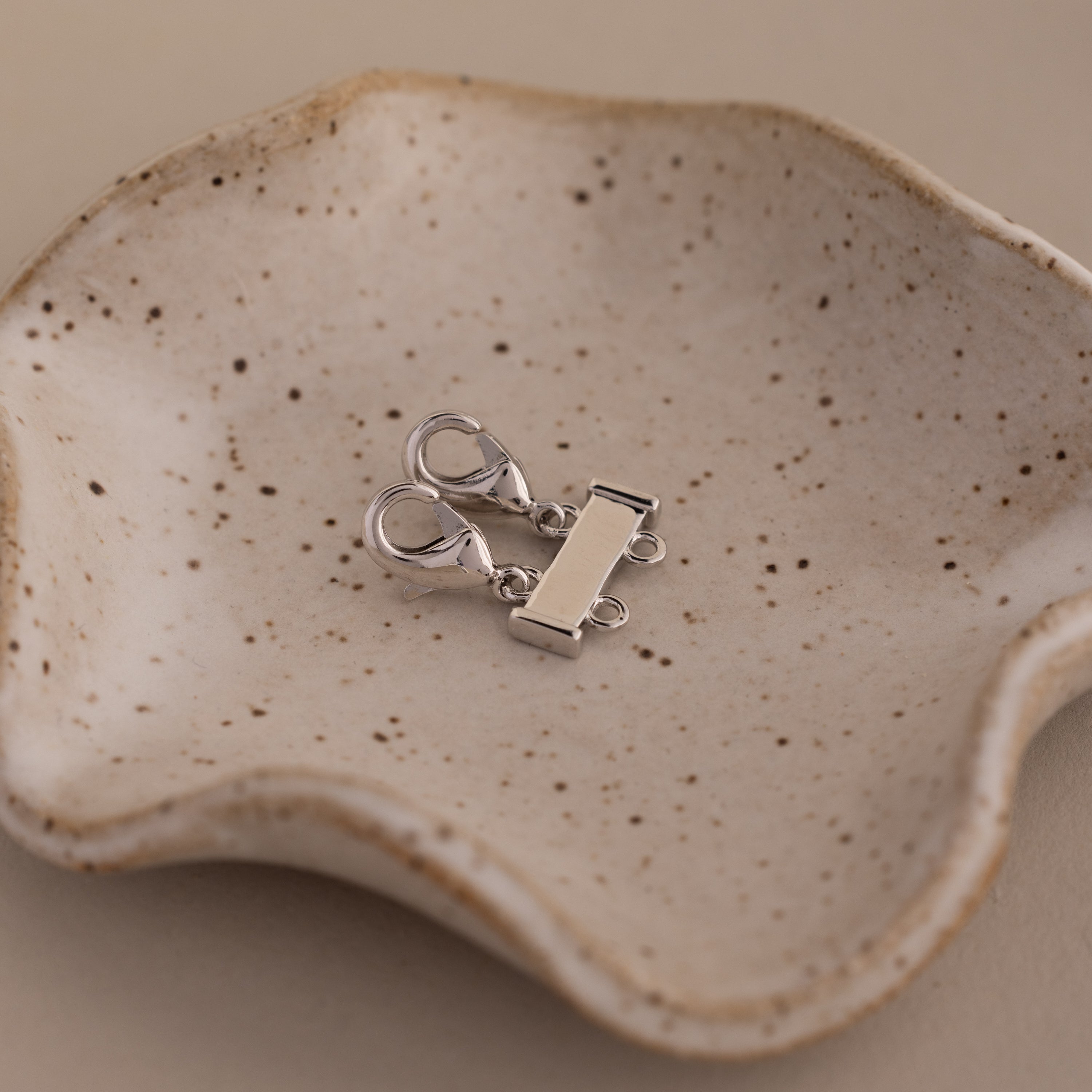 The Double Layering Clasp in Silver rests on a speckled ceramic dish with wavy edges, perfect for layering necklaces, set against a beige background.