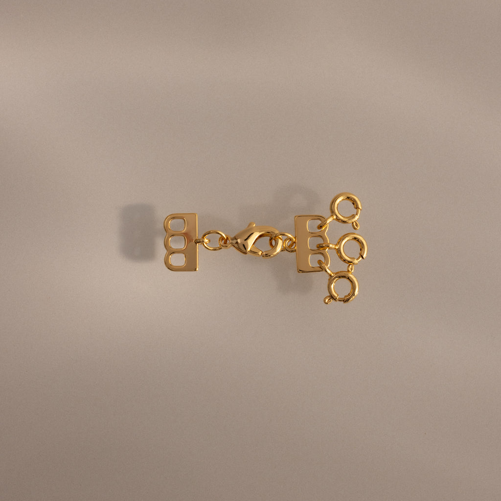 The Triple Layering Clasp in 18K Gold features three loops—ideal for organizing your gold and silver necklaces or creating a chic layered look on any neutral background.