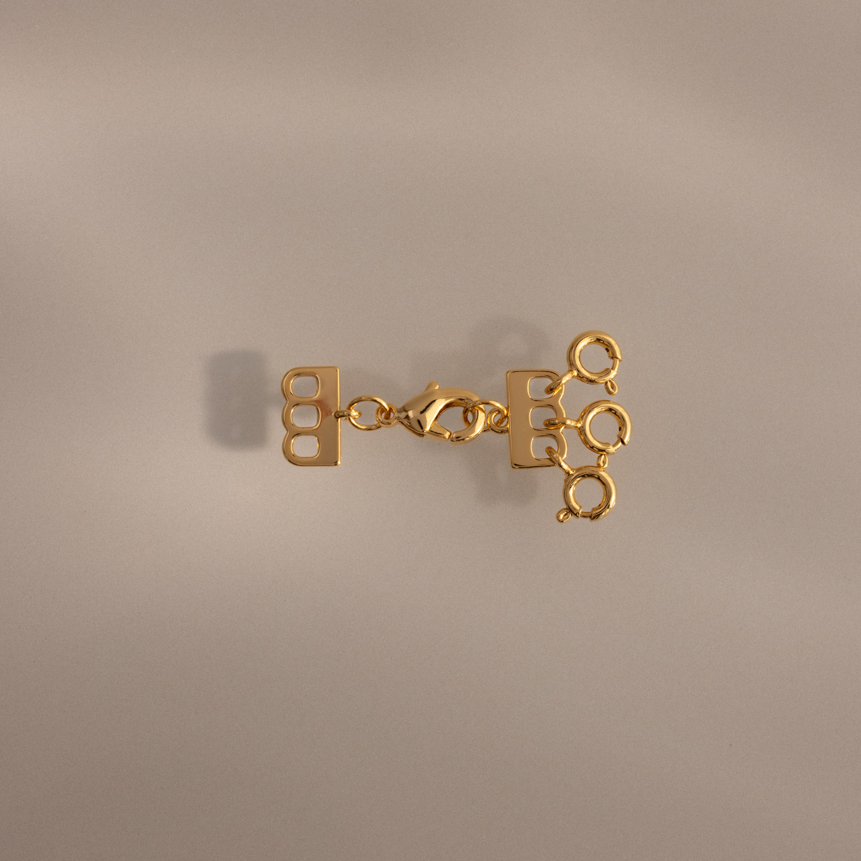 The Triple Layering Clasp in 18K Gold features three loops—ideal for organizing your gold and silver necklaces or creating a chic layered look on any neutral background.