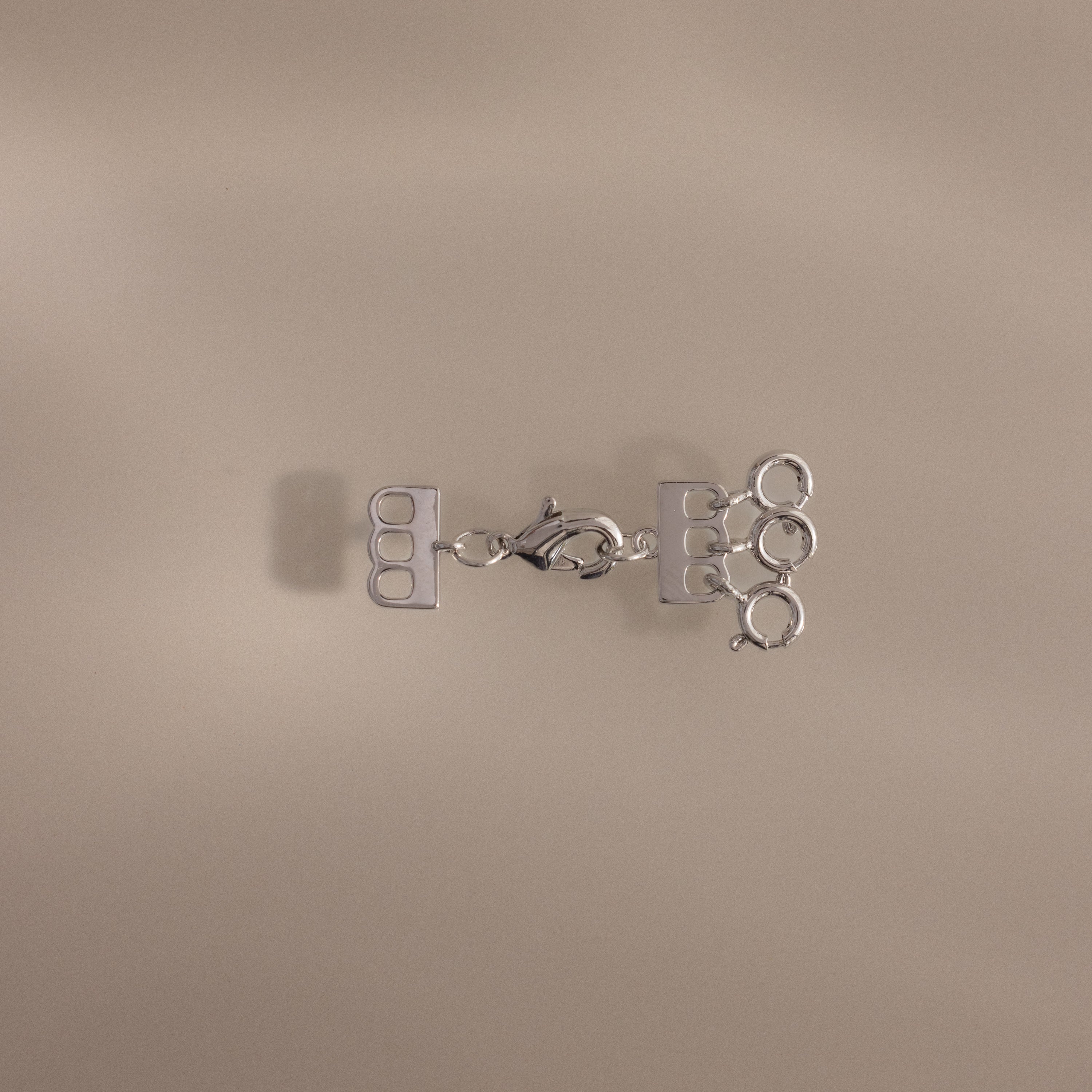 The Triple Layering Clasp in Silver features three loops and a clasp for tangle-free necklace layering, shown on a beige background.