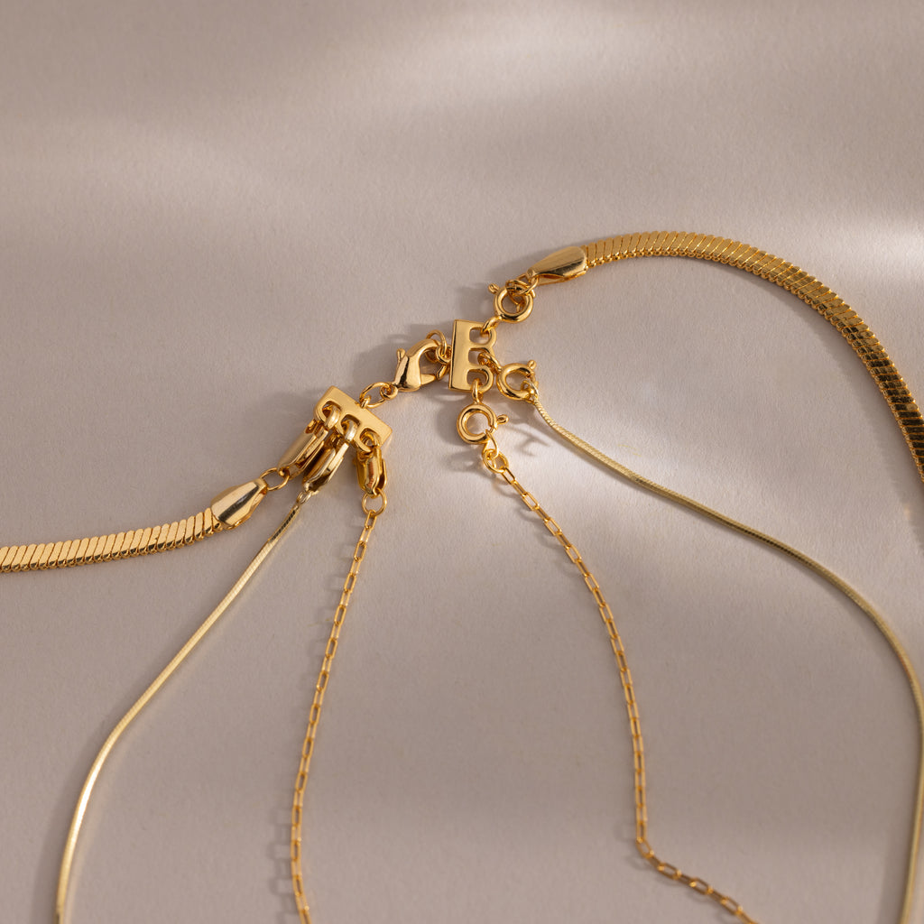 Four gold necklaces with different chain styles are displayed on a neutral background, highlighting the Triple Layering Clasp for easy necklace stacking and tangle-free wear.