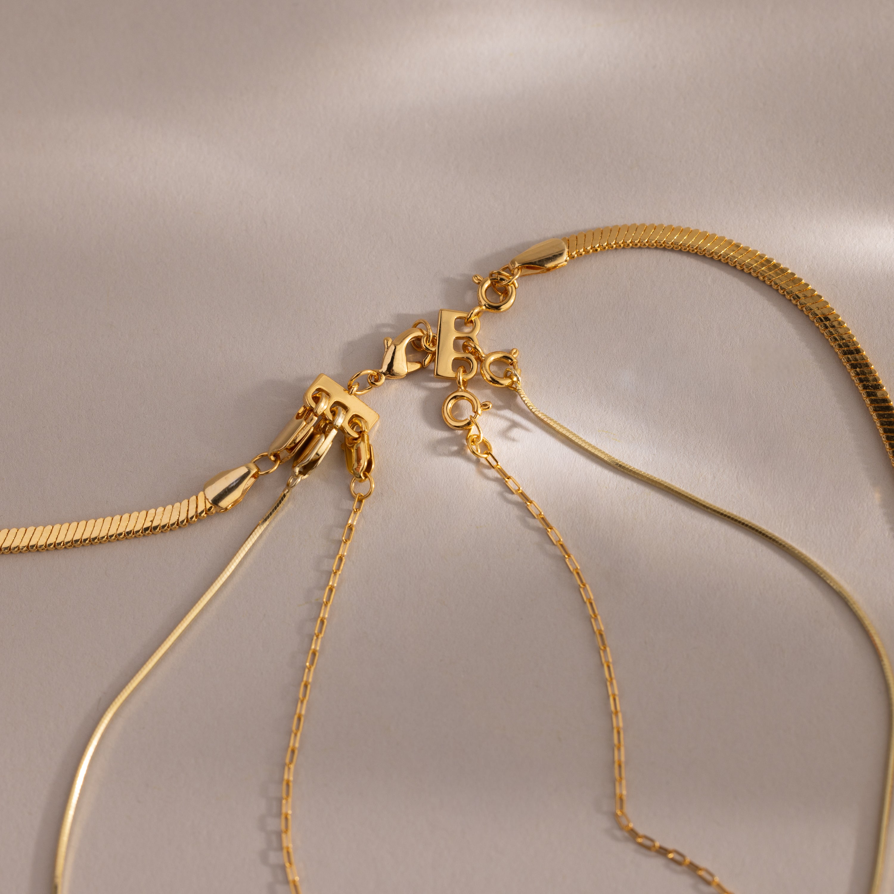 Four gold necklaces with different chain styles are displayed on a neutral background, highlighting the Triple Layering Clasp for easy necklace stacking and tangle-free wear.