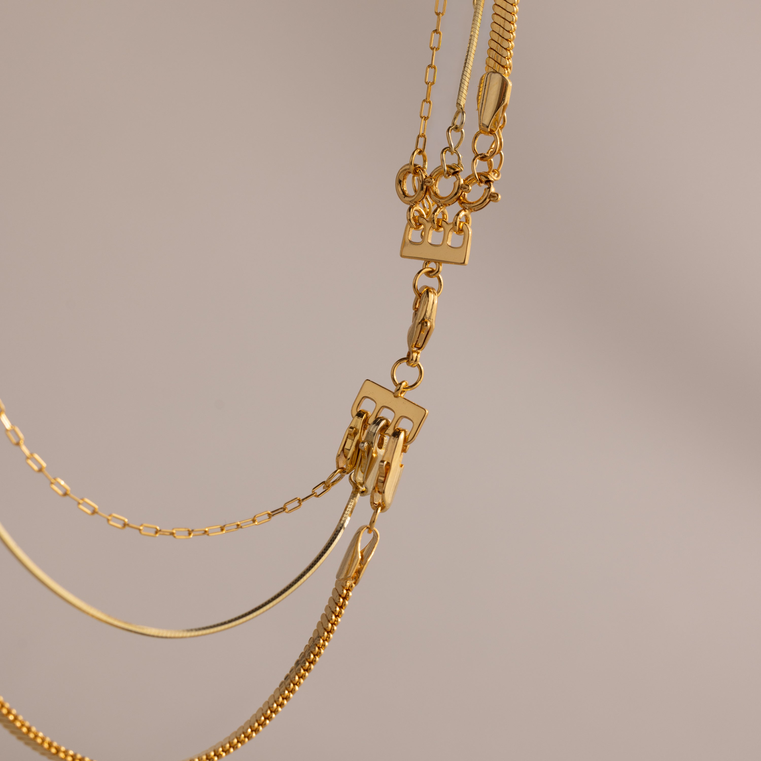 Close-up of layered gold necklaces with various chain styles, highlighted by the Triple Layering Clasp in 18K Gold for easy necklace layering, set against a neutral background.
