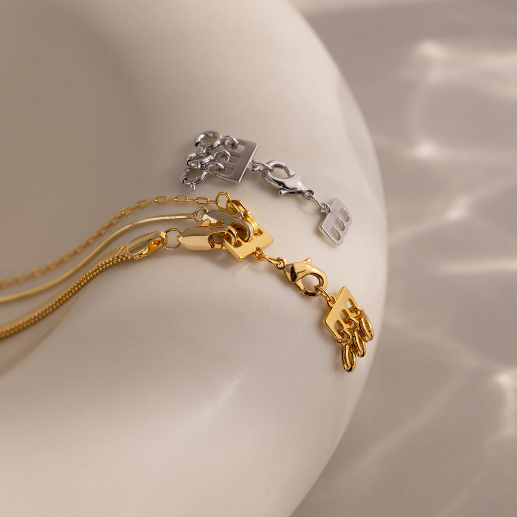 The Triple Layering Clasp keeps gold and silver chain necklaces with “bb” letter pendants tangle-free, perfect for stacking and featuring secure lobster claw clasps on a smooth beige surface.
