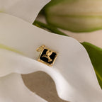 The Onyx Hummingbird Charm in 18K Gold rests on soft white petals and green leaves—a standout jewelry piece ideal for anyone seeking a nature-inspired look.