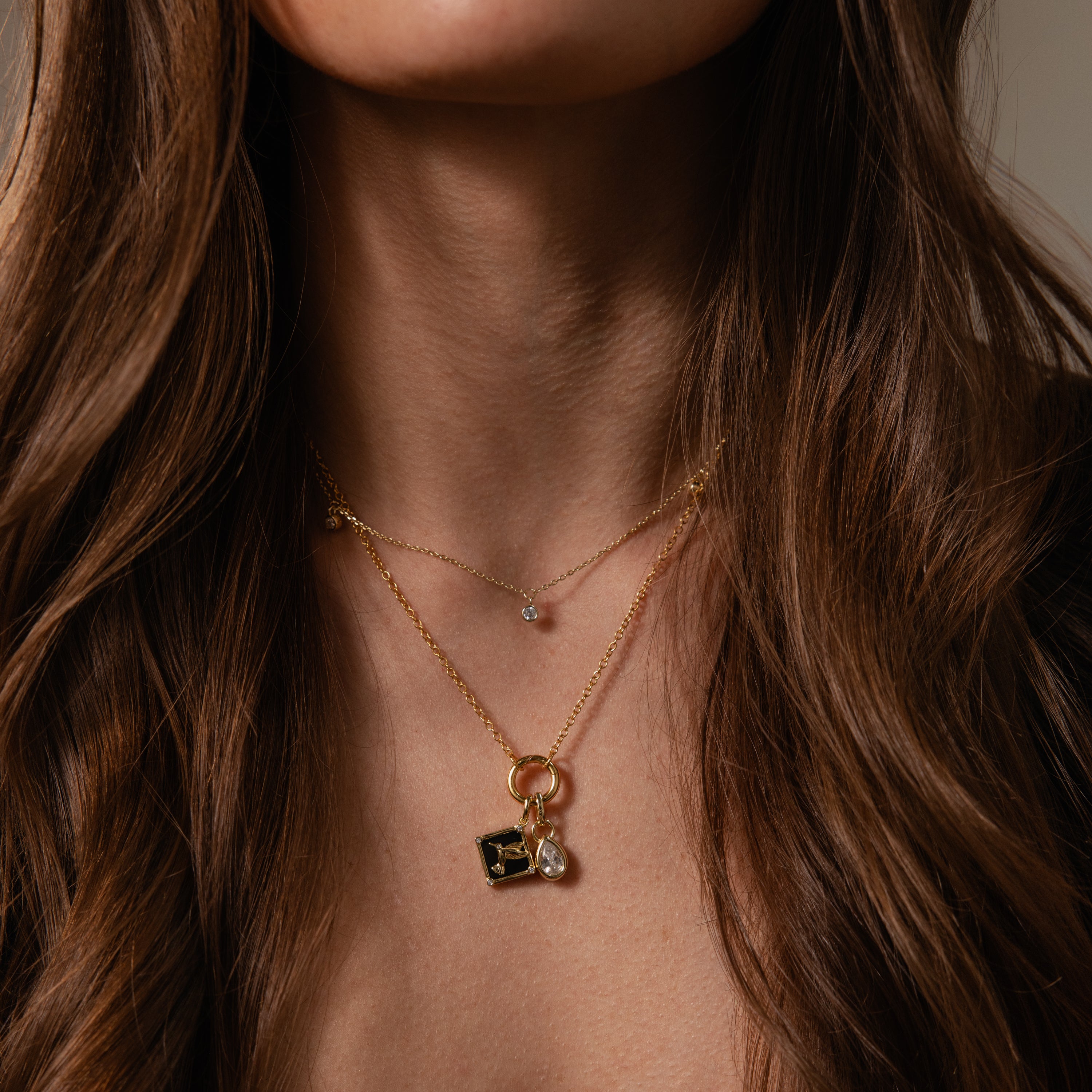 Woman with long brown hair wears the Onyx Hummingbird Charm in 18K Gold, layered with other gold necklaces for a delicate, elegant look perfect for everyday wear.