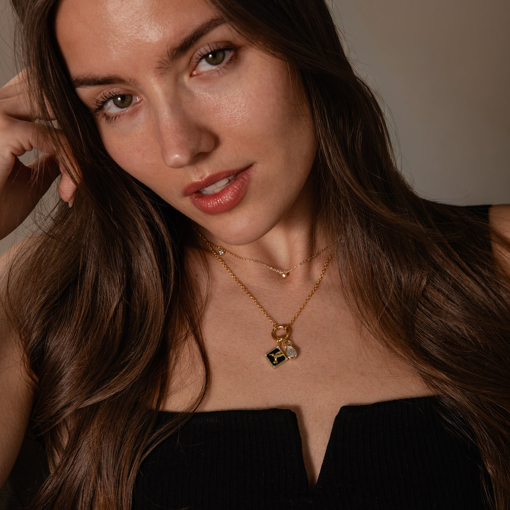 Woman with long brown hair wears a black top and layered gold necklaces, featuring the Onyx Hummingbird Charm, looking at the camera.