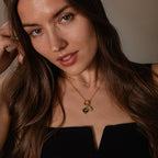 Woman with long brown hair wears a black top and layered gold necklaces, featuring the Onyx Hummingbird Charm, looking at the camera.