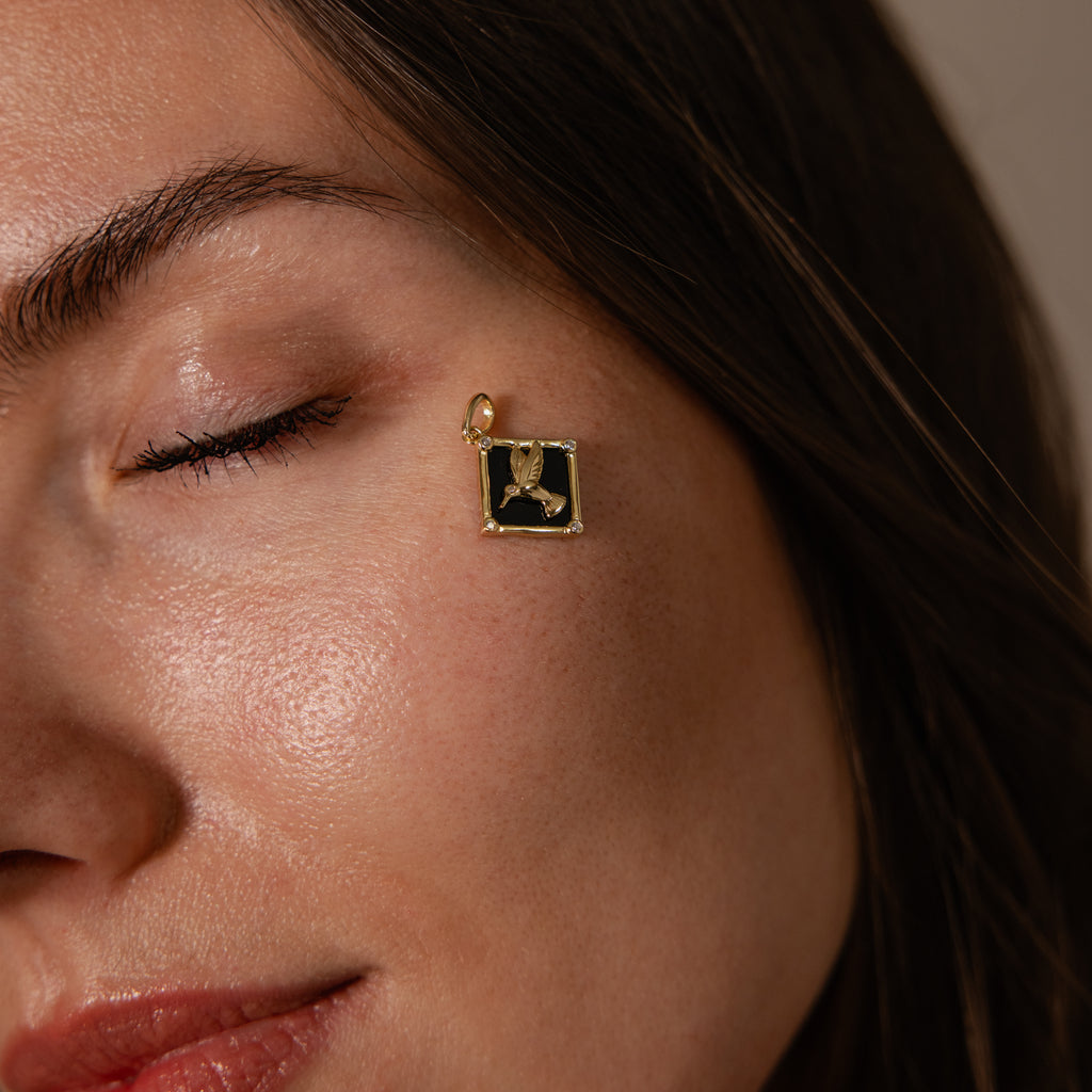 A woman with closed eyes showcases the Onyx Hummingbird Charm in 18K Gold, wearing it as an elegant pendant earring near her eye to highlight its refined style and unique jewelry details.