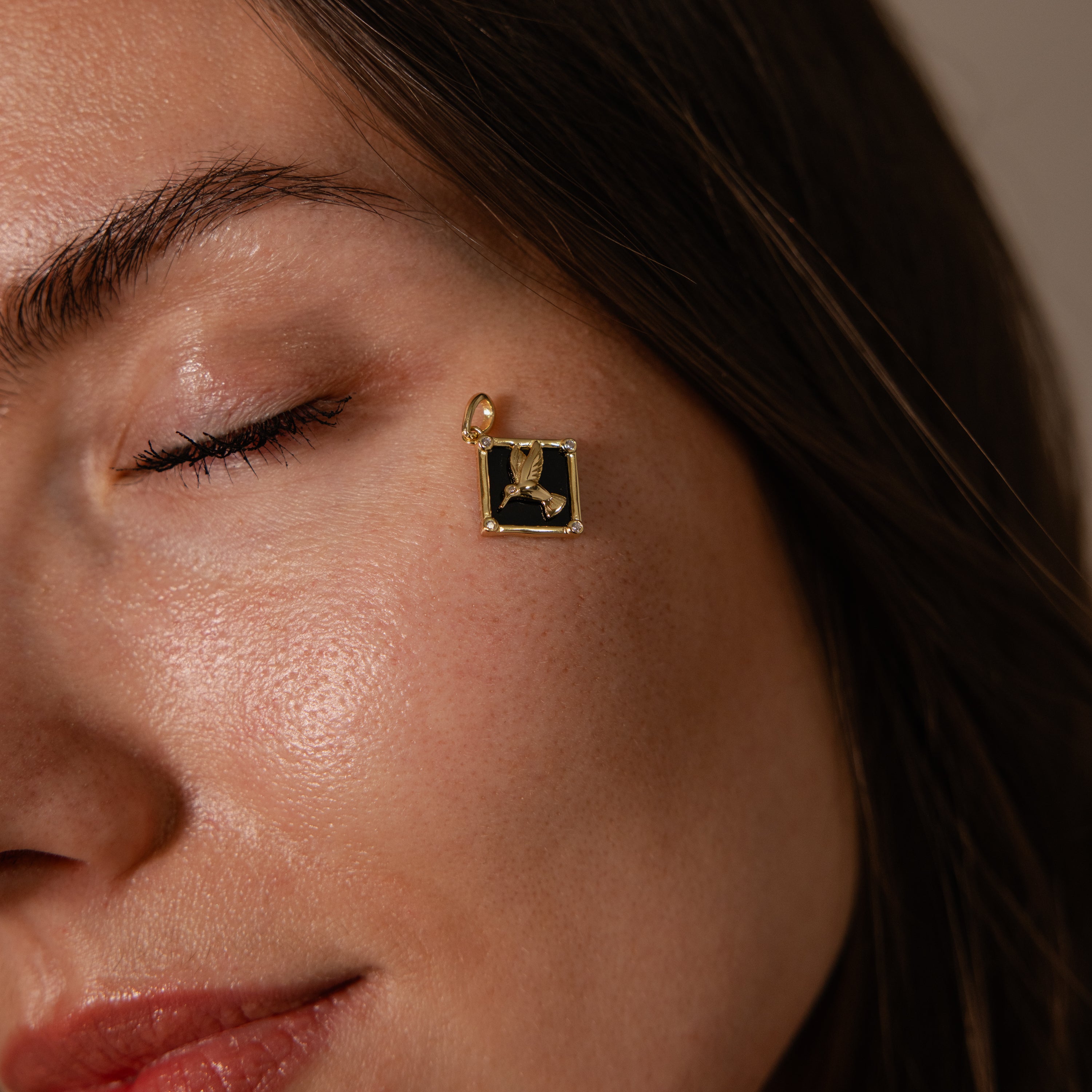 A woman with closed eyes showcases the Onyx Hummingbird Charm in 18K Gold, wearing it as an elegant pendant earring near her eye to highlight its refined style and unique jewelry details.